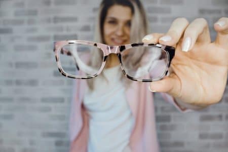 Les symptômes qu’il est temps pour vous de mettre des lunettes Young woman with blonde hair holding eyeglasses indoors, focusing on vision aid.