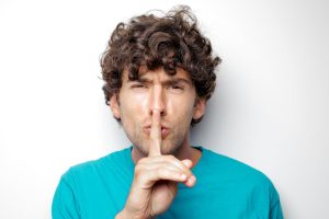 Adult man with curly hair in blue shirt making a silence gesture on white background.