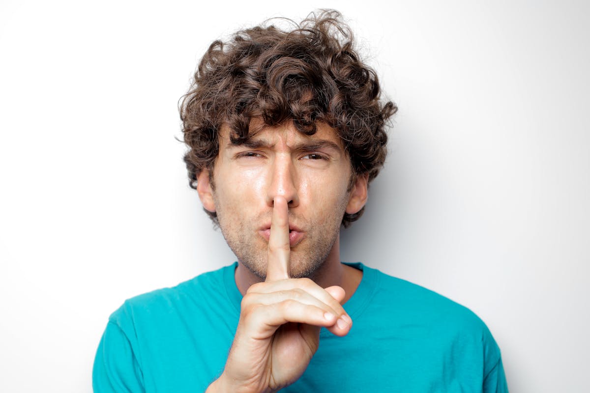 Le Canard enchaîné révèle les clauses de silence imposées aux journalistes quittant les médias Bolloré Adult man with curly hair in blue shirt making a silence gesture on white background.