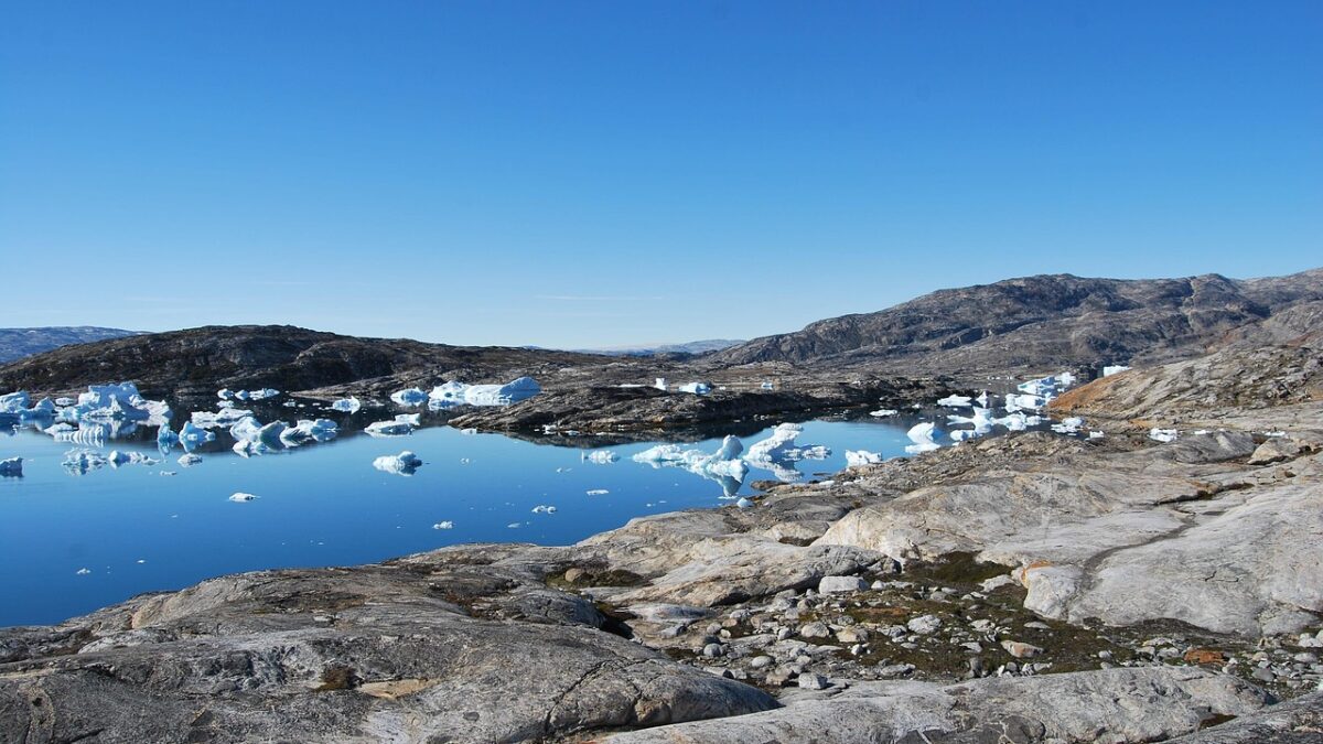 Paysage rocheux au bord d'une étendue d'eau bleue calme, parsemée de petits icebergs blancs, sous un ciel bleu dégagé, avec des collines en arrière-plan.