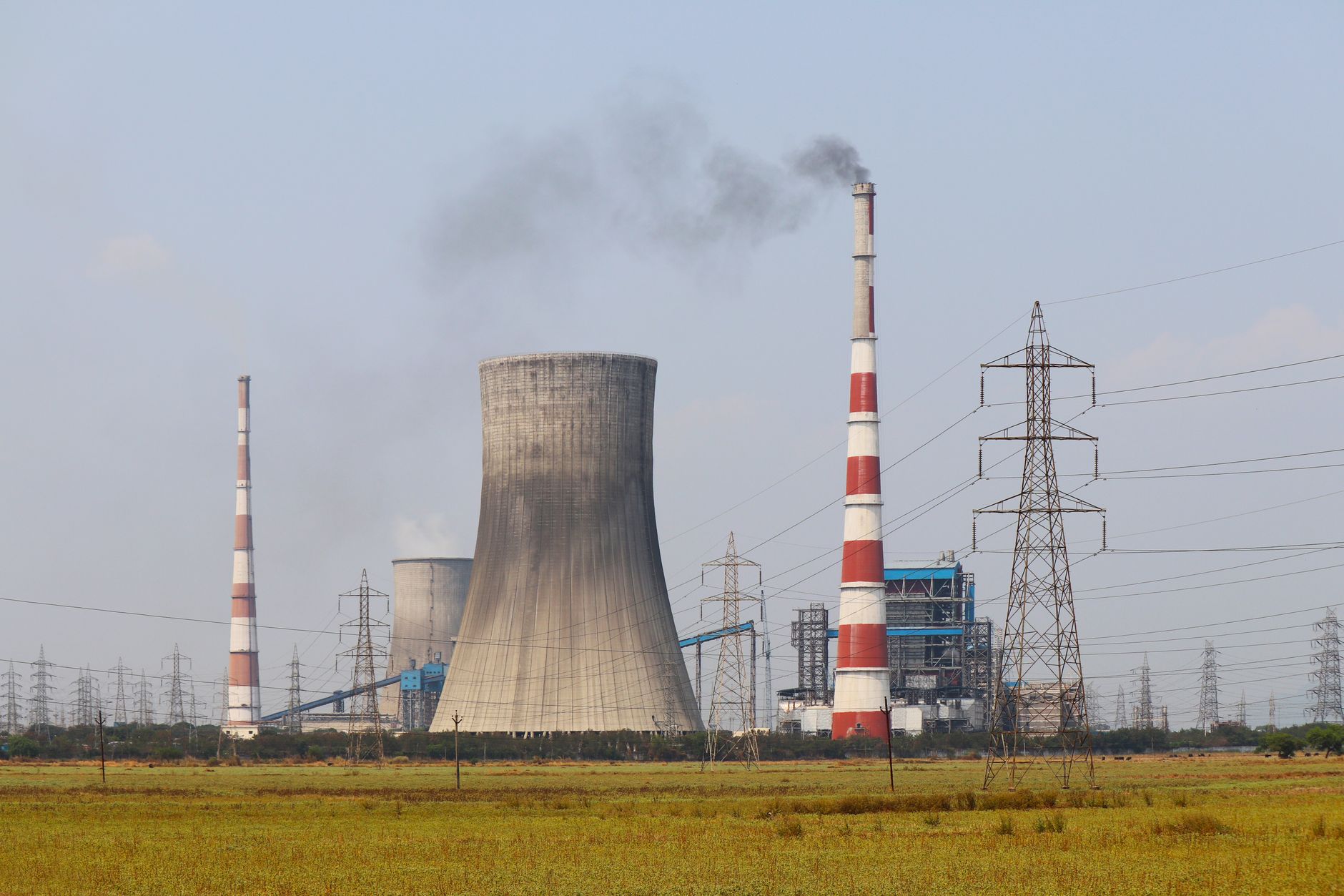 Centrale thermique avec tour de refroidissement, cheminées rouge et blanc dont une fume, et pylônes électriques derrière un champ.
