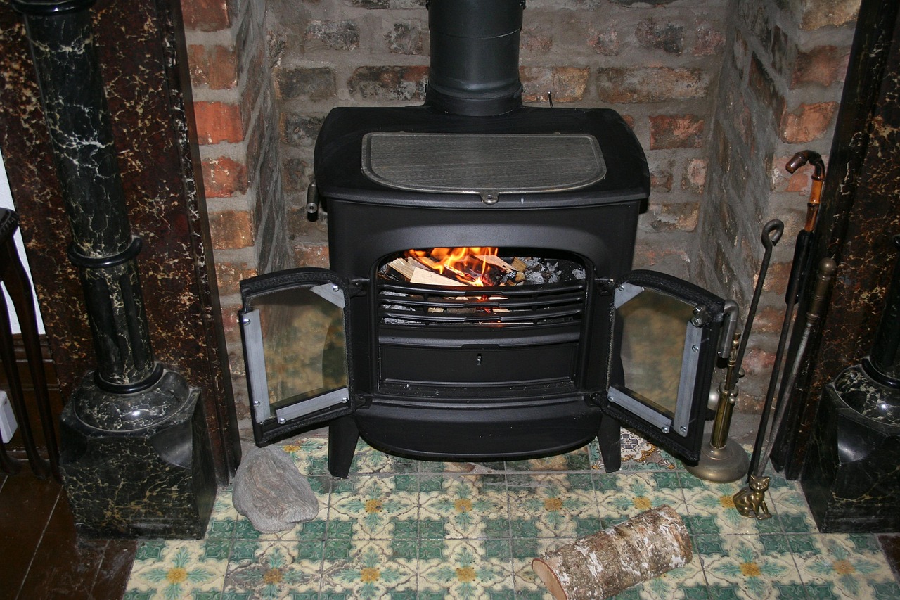 Poêle à bois noir aux portes ouvertes, avec des flammes et des bûches à l'intérieur, devant un mur de briques et un sol de carreaux colorés