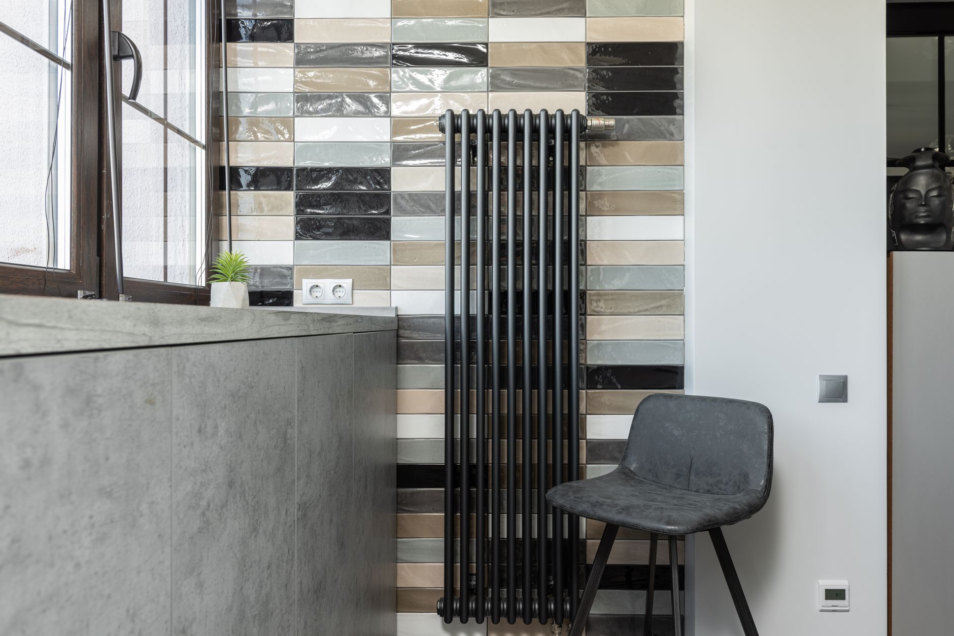 Radiateur vertical noir à colonnes sur un mur de carrelage multicolore, avec une chaise grise et un plan de travail gris.